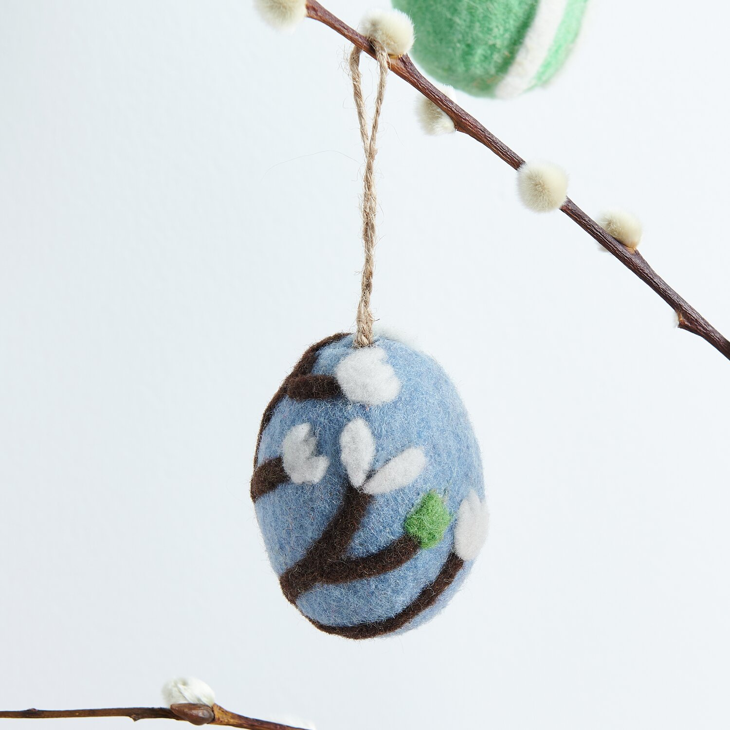 Aufhänger Filz-Ei Kirschblüte: Blaues Filz-Ei mit Kirschblütenmuster, hängend an einem Ast. Größe 7,8xØ5,8cm.