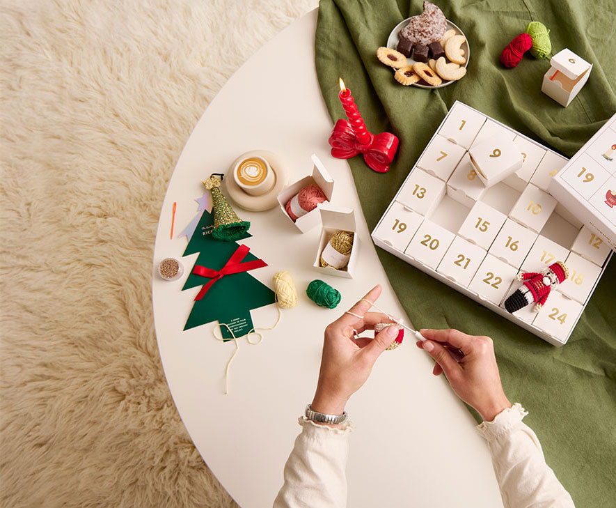 Ricorumi Adventskalender auf Tisch Ricorumi Adventskalender liegt geöffnet auf einem Tisch und Person häkelt.