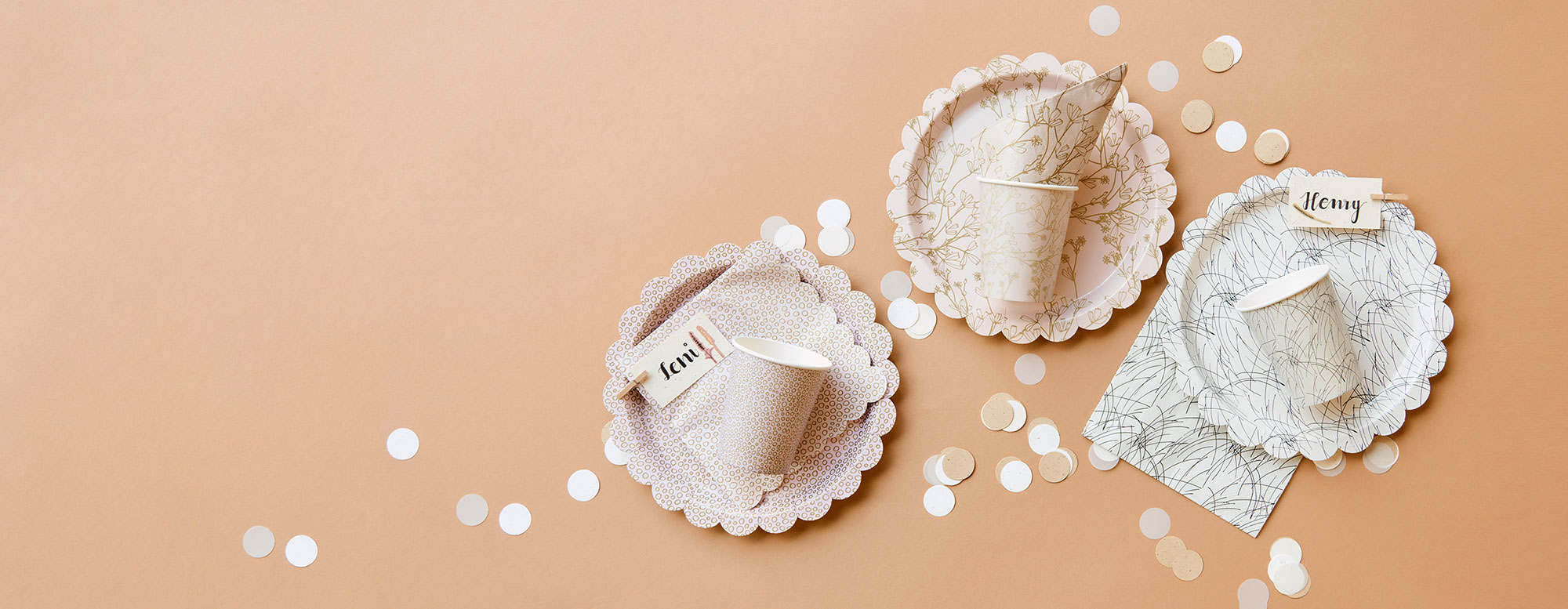 Pappteller, -becher und Servietten in drei unterschiedlichen Motiven, dazu bräunliches Konfetti. Flatlay von Produkten aus der Kollektion In The Meadow.