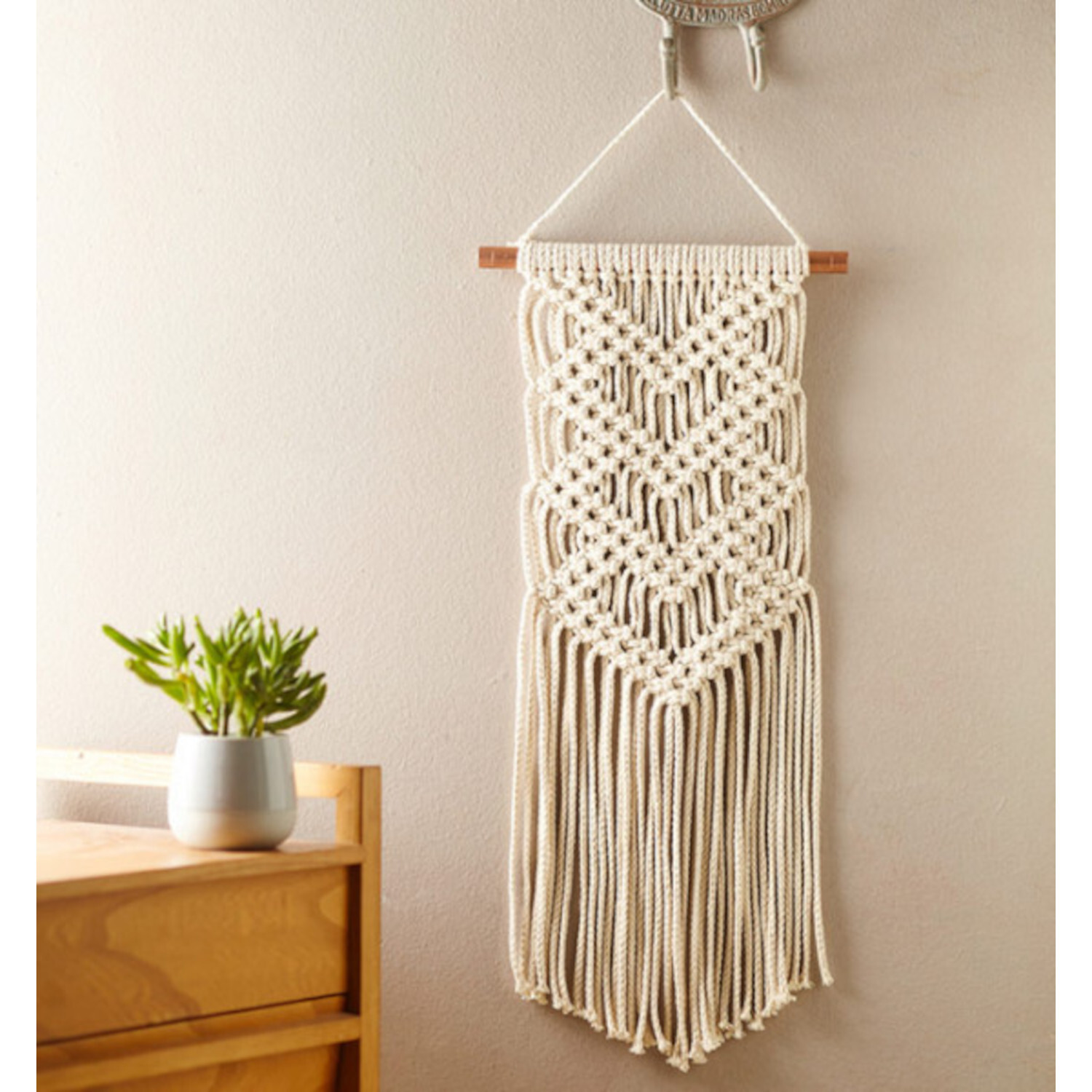 Makramee für Anfänger - Step-by-Step Einsteigerbuch: Wandbehang aus cremefarbenem Garn, dekorativ an einer Holzstange aufgehängt.