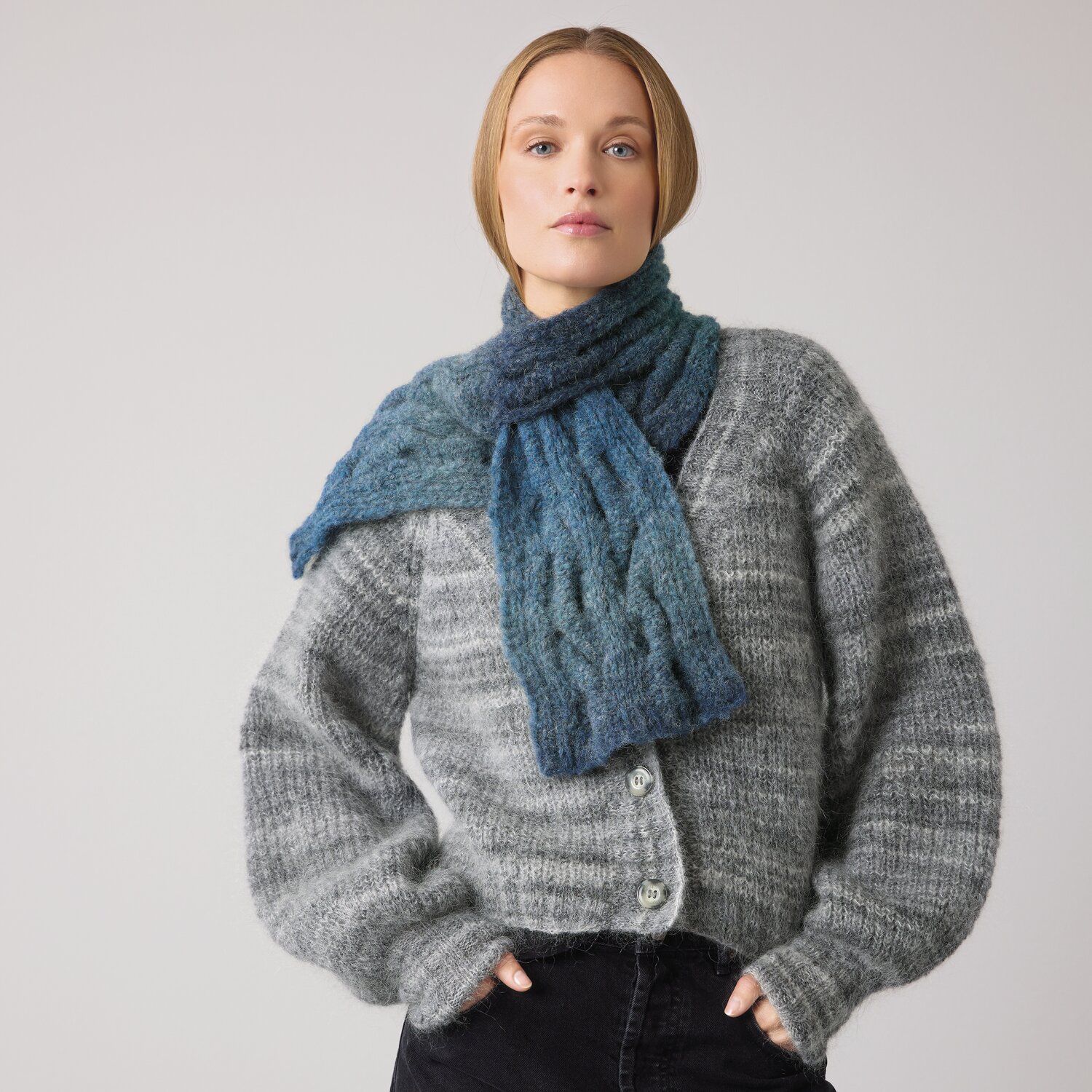 Strickset Schal Modell 16 aus Die Neue Masche No. 14 von idee. Creativmarkt Strickset Schal Modell 16 aus DNM 14: Frau trägt blauen Schal und graue Strickjacke vor grauem Hintergrund.