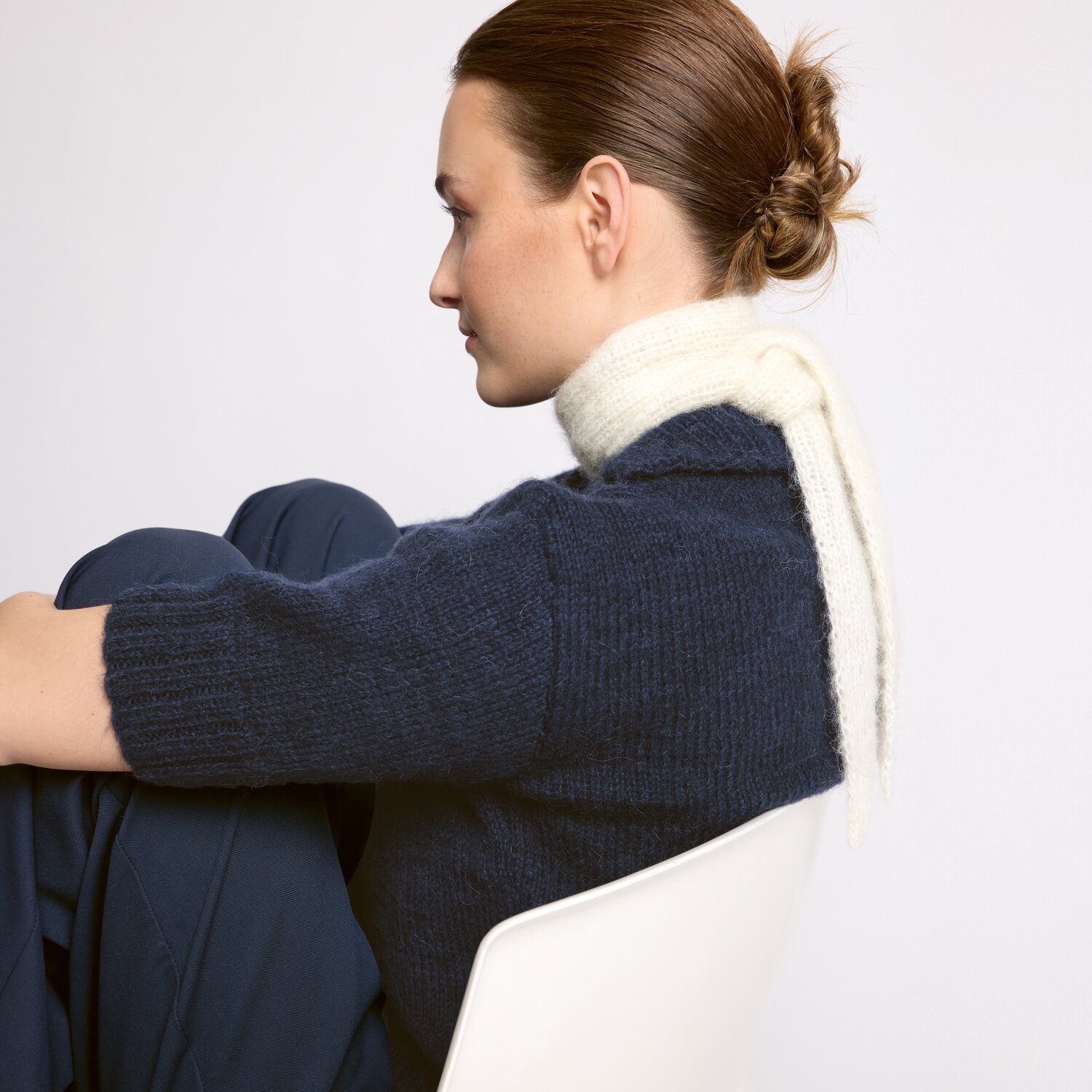 Strickset Tuch Modell 08 aus Luxury Knits - Premium Natural Fibres von Rico Design Strickset Tuch Modell 08 aus Luxury Knits: Frau mit weißem Schal und dunkelblauem Pullover sitzt seitlich auf einem Stuhl.