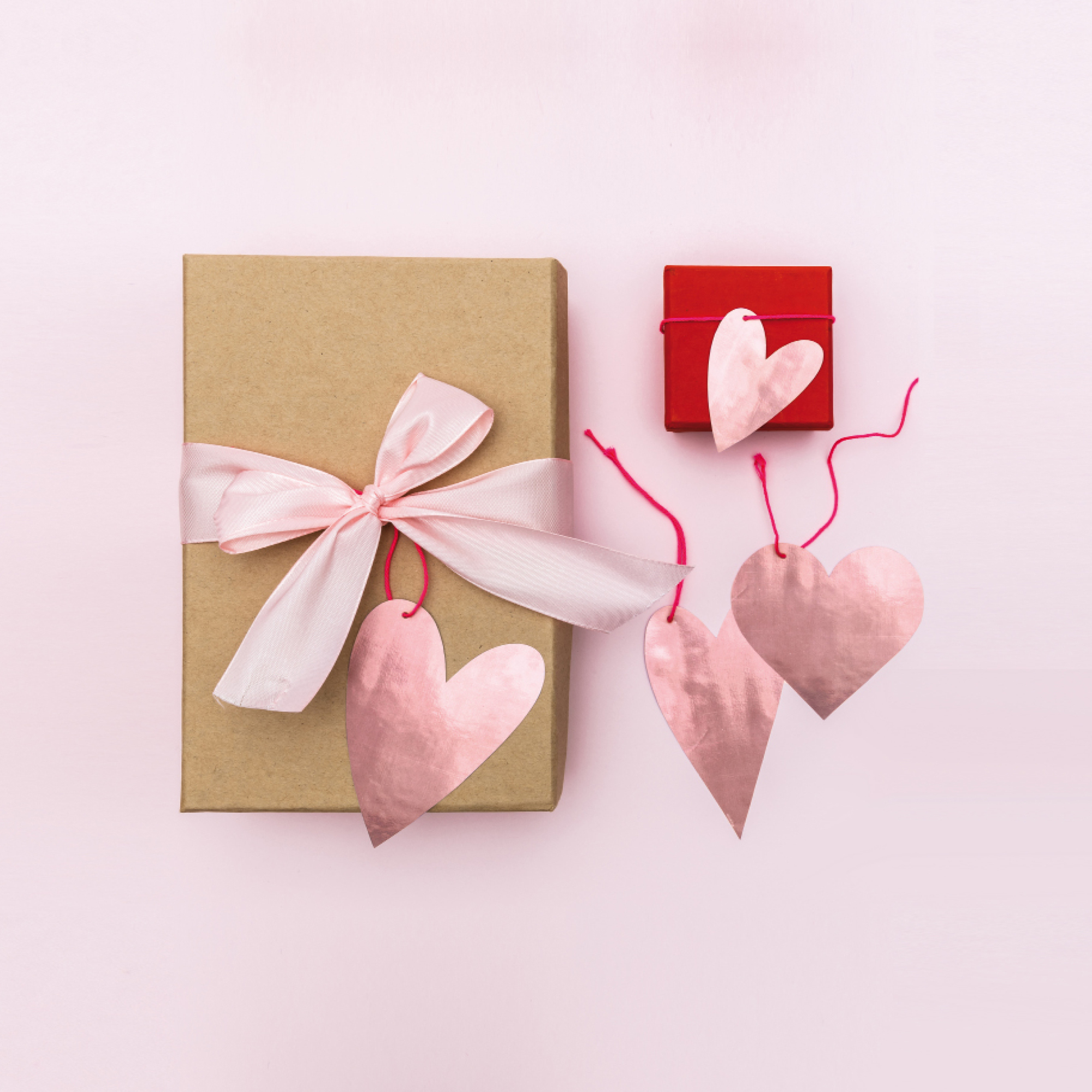 Verpacktes Geschenk mit rosa Schleife und Herz-Anhänger und kleines verpacktes Geschenk mit kleinem Herz-Anhänger. Flatlay von Produkten.