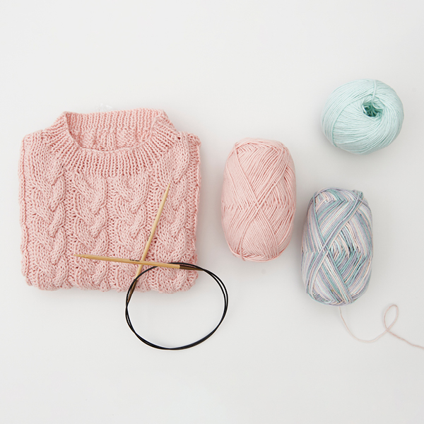 Ein gestrickter und gefalteter Pullover aus rosa Garn, dazu Rundstricknadel und drei Garnknäule. Flatlay von Produkten.