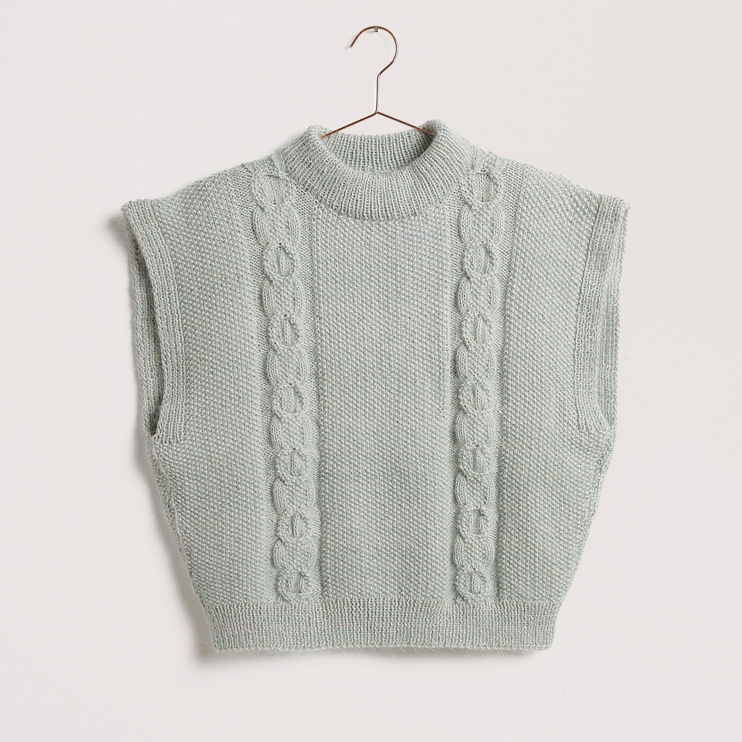 Die Neue Masche No. 12 Frühjahr-Sommer von idee. Creativmarkt Strickweste in Silbergrau mit Zopfmuster, ärmellos und rundem Kragen, auf einem Kleiderbügel.