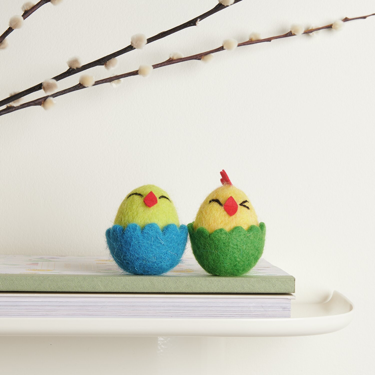 Filz-Küken im Ei Gelb/Blau und Gelb/Grün auf einem Tisch, dekorativ platziert vor einem Zweig.