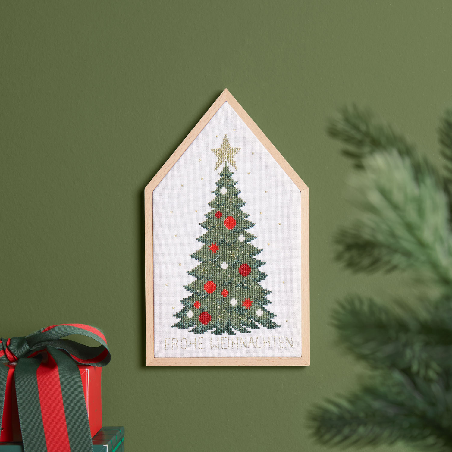 Ein Holzstickrahmen mit einem Weihnachtsbaum bestickt hängt an einer grünen Wand.