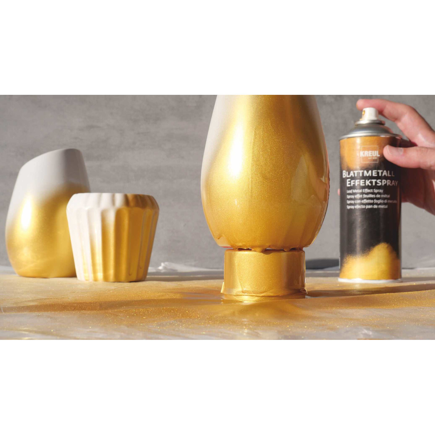 Blattmetall Effektspray verleiht Keramikvasen und -gefäßen einen goldenen Glanz. KREUL Spraydose in Aktion auf grauem Hintergrund.