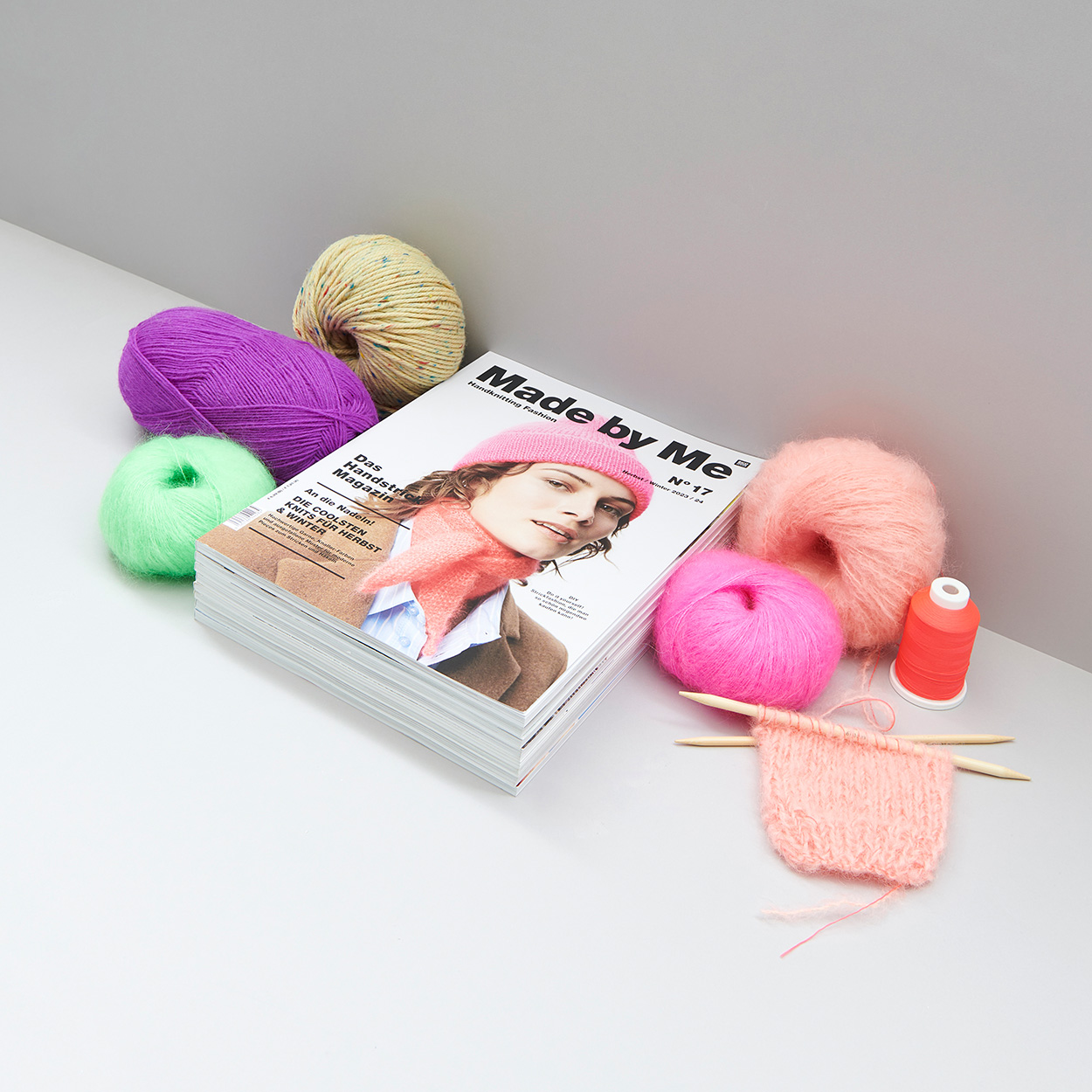 Stapel von Zeitschriften daneben Garne und ein Strickstück mit Stricknadeln. Fotosetting graue Ecke.