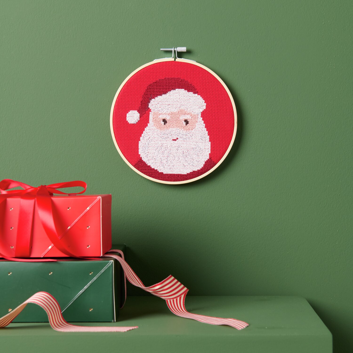 Stickpackung Portrait Santa Ø15cm von Rico Design Stickpackung Portrait Santa zeigt ein gesticktes Weihnachtsmann-Gesicht auf rotem Hintergrund, aufgehängt an einer grünen Wand.
