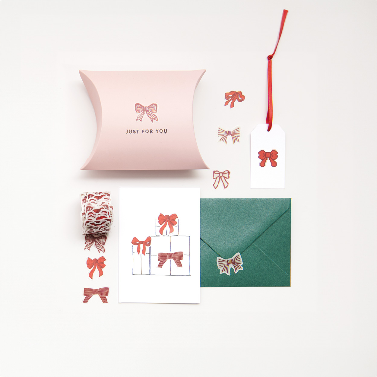 Rosa Geschenkschachtel, weiße Karte, grüner Umschlag und weißer Geschenkanhänger mit roten und rosa Schleifensticker verziert, sowie Rolle mit Schleifensticker.