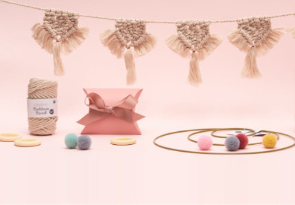 Hängende Makramee-Girlande mit Garn, eine Geschenkschachtel, verschiedene Dekoringe und Verpackung mit Garn-Pompons. Fotosetting rosa Wand.