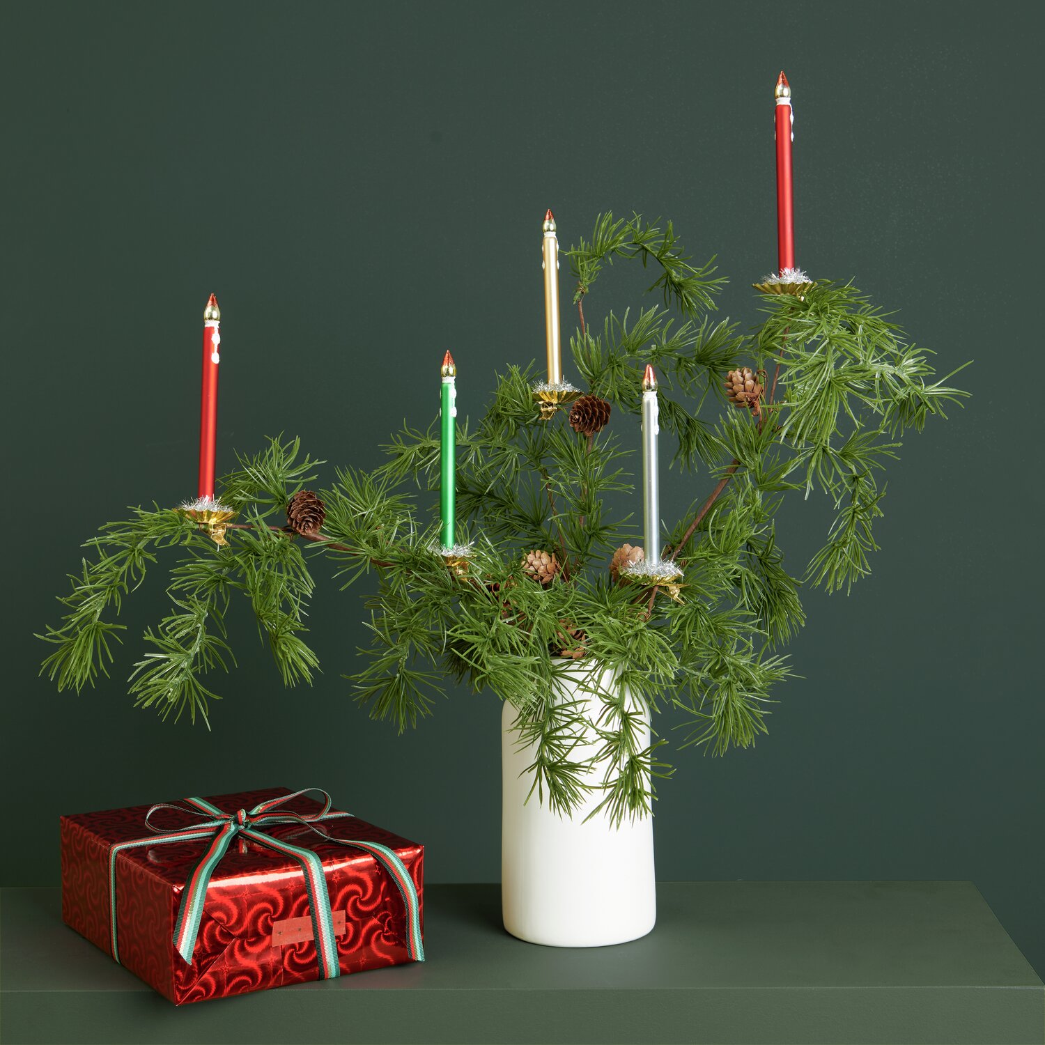 Baumschmuck Glaskerze mit goldenem Metallchip auf Tannenzweigen, umgeben von roten Geschenken. Farben: Gold, Grün, Silber, Rot.