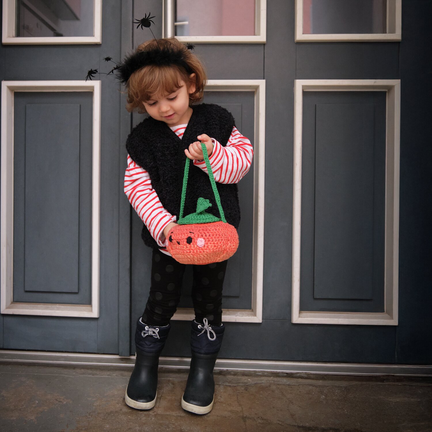 Haarreif Spinnen schwarz: Kind mit schwarzem Spinnen-Haarreif und Kürbistasche vor blauer Tür.