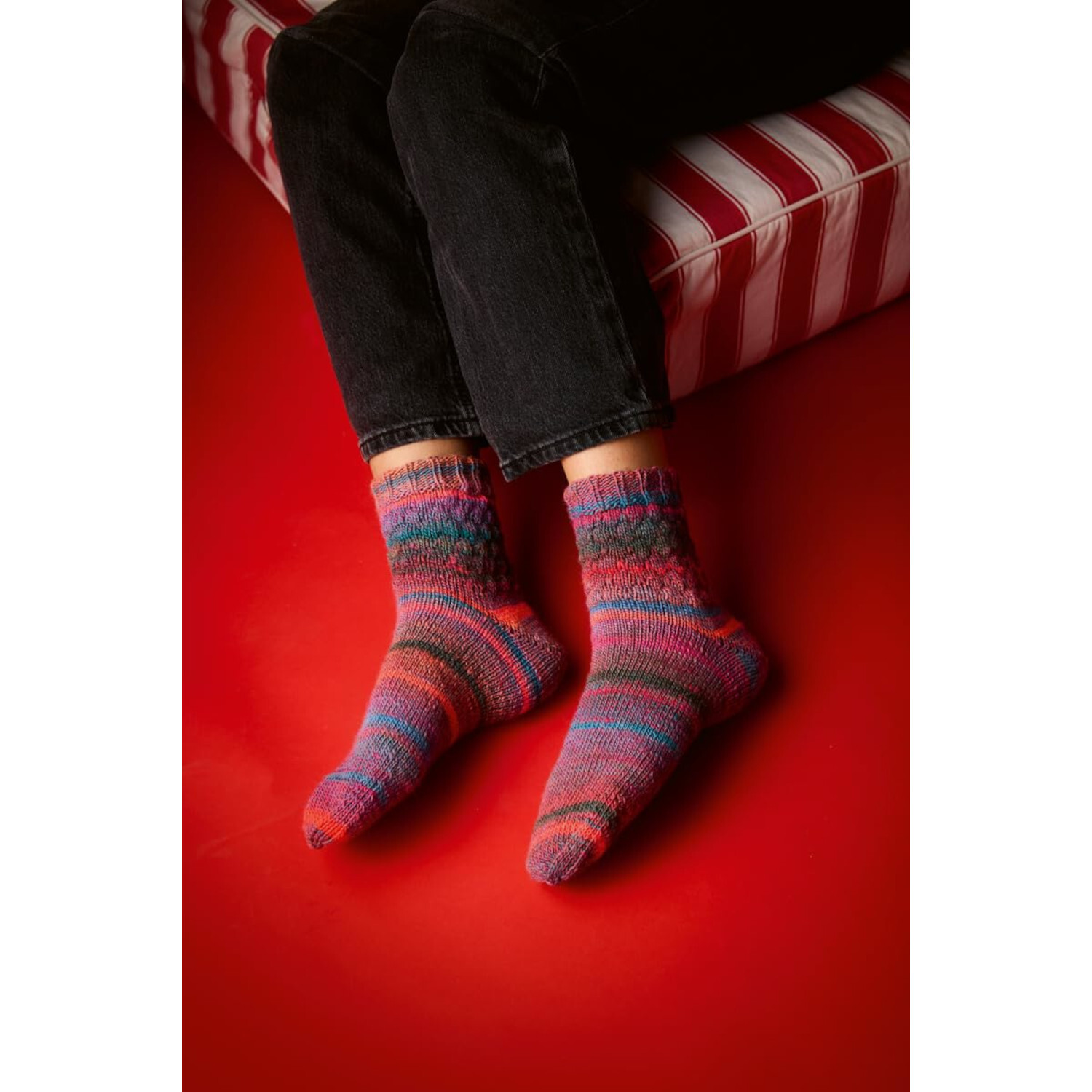 Socken stricken mit Wow-Effekt: Bunte, handgestrickte Socken auf rotem Boden, getragen von Person in schwarzer Hose.