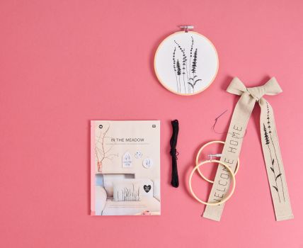 Stickset mit Stickrahmen, Stickgarn, Stoffschleife und Anleitung auf rosafarbenem Hintergrund, verziert mit filigranem Wiesenblumenmotiv.
