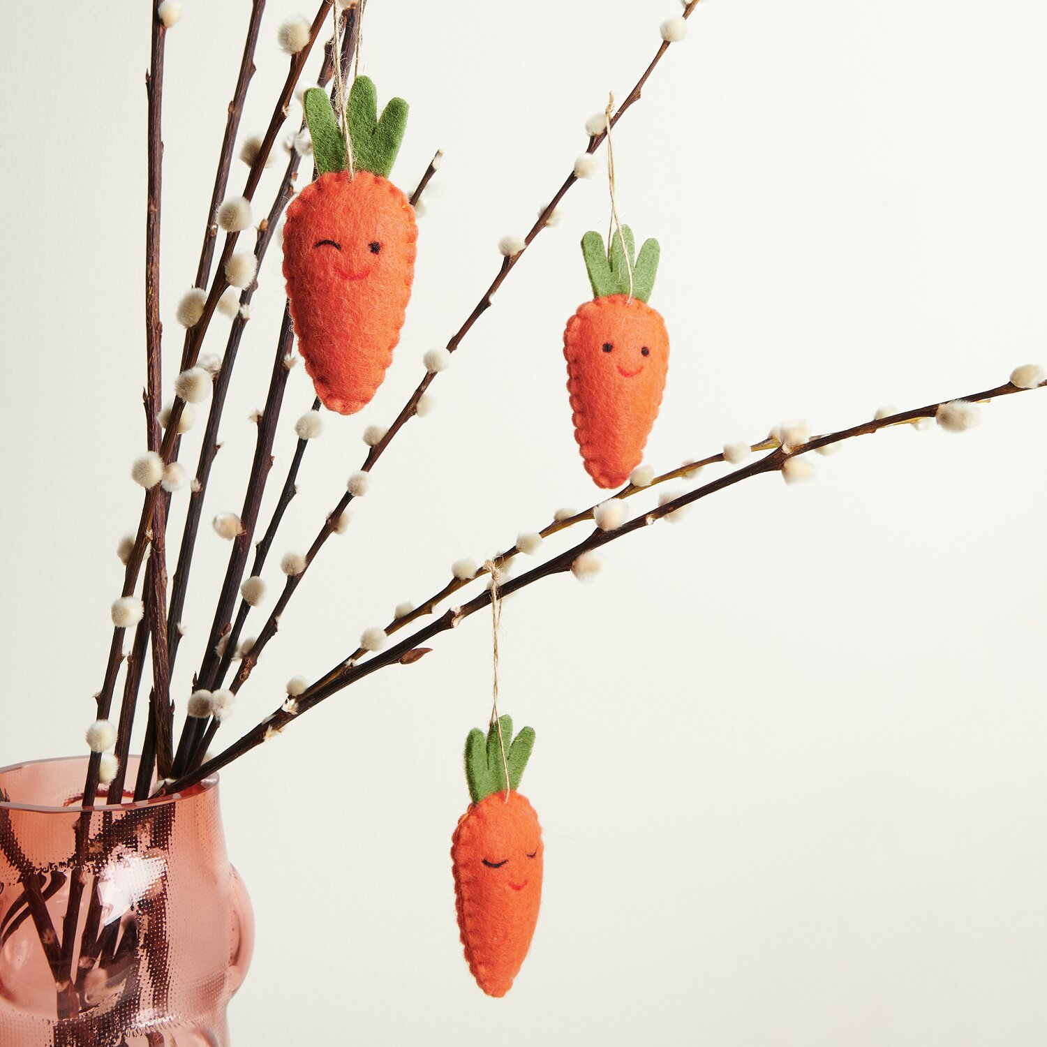 Aufhänger Filz-Möhre Zwinkernd 2D: Drei orange Filz-Möhren mit Gesichtern hängen an Zweigen in einer Vase.