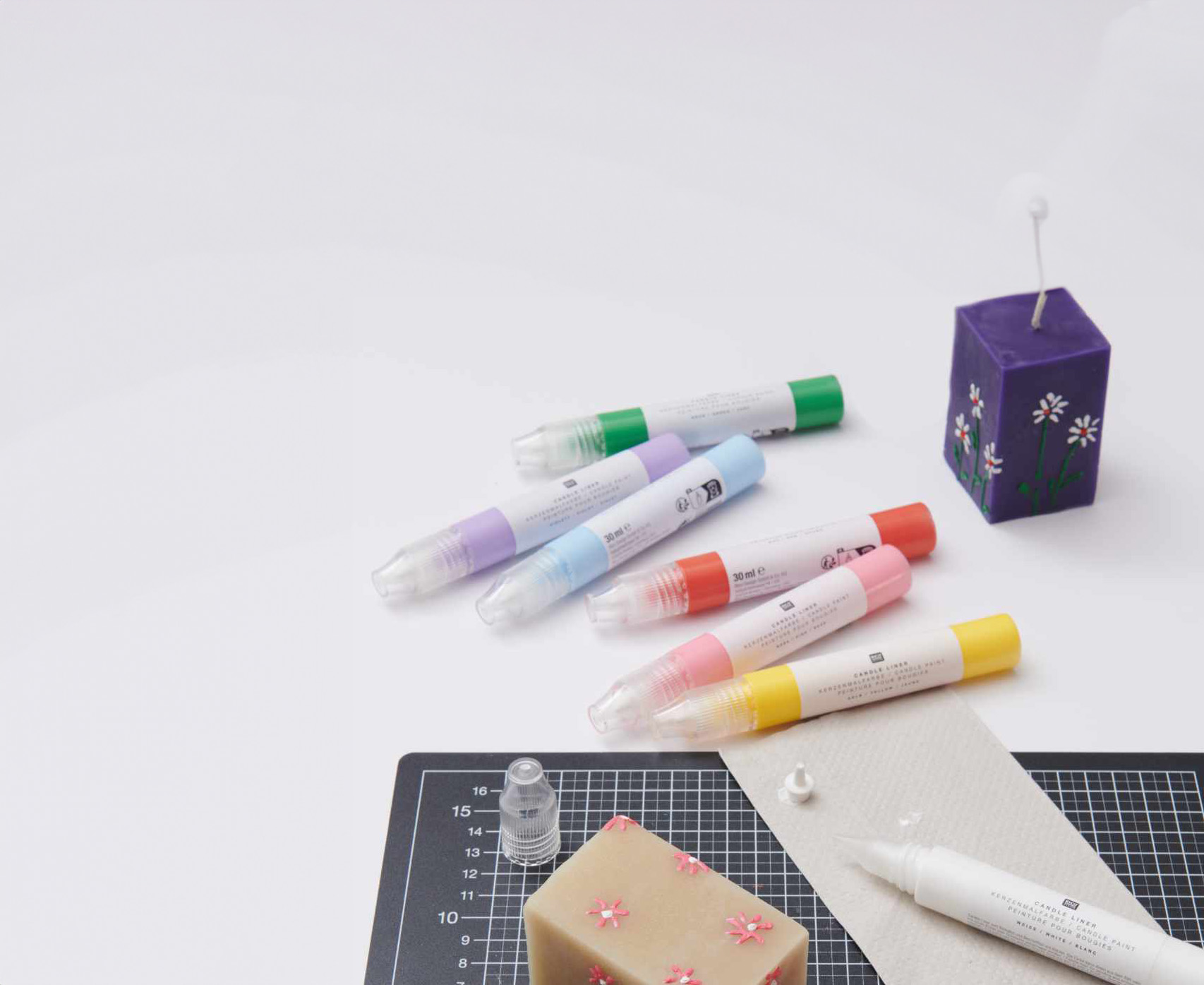 Eine lila und eine beige eckige Kerze mit Blumendesign, daneben verschieden farbigen Candle Liner. Flatlay von Produkten.