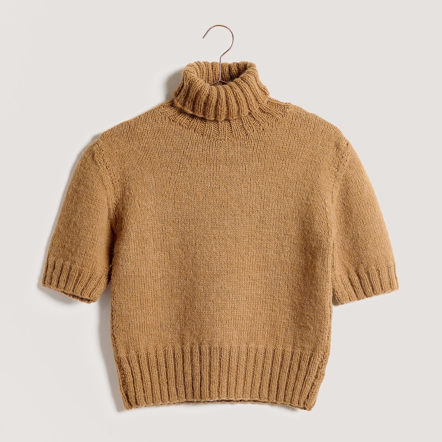 Strickset Shirt Modell 01 aus Lovewool No. 23, kurzärmliger Rollkragenpullover in warmem Camelton, locker gestrickt, gerippte Bündchen, auf Bügel vor hellem Hintergrund.