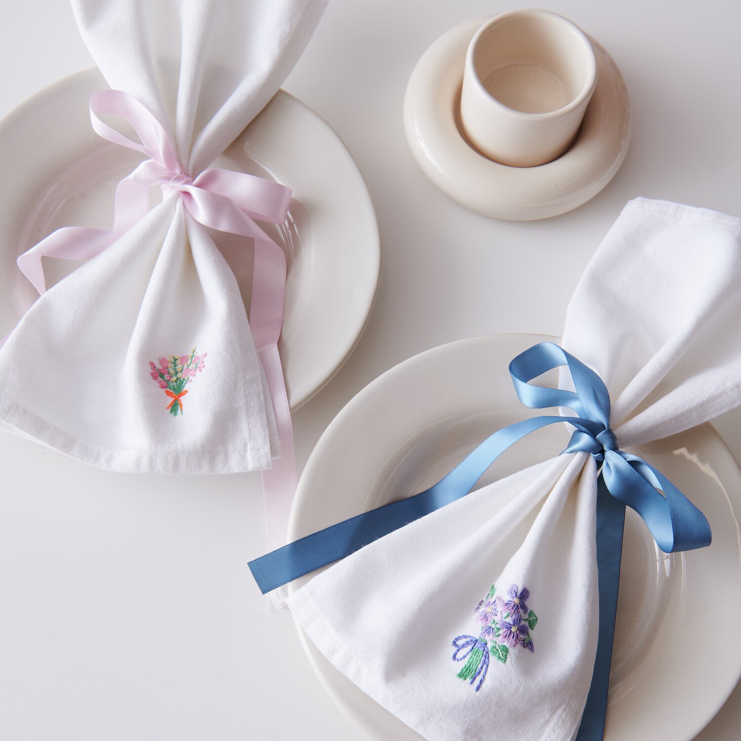 Stick & Stitch Packung Blumensträuße XL 7 Stück: Weiße Servietten mit Blumenstickerei, rosa und blaue Schleifen, auf weißen Tellern arrangiert.