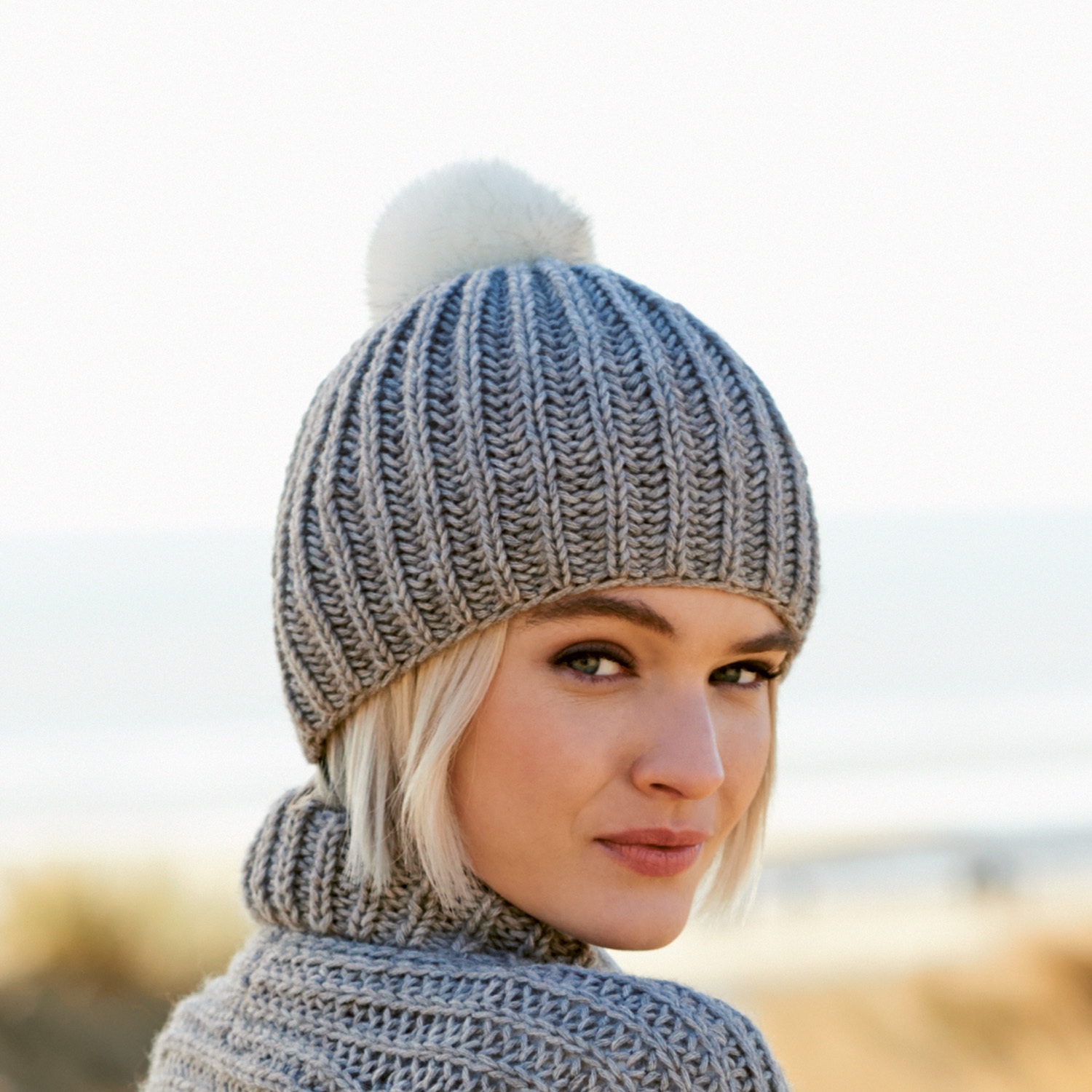 Strickanleitung Mütze mit Kunstfellpompon: Graue Strickmütze mit weißem Pompon, getragen von Frau am Strand.