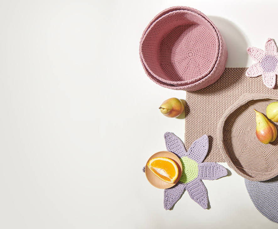 Drei pinke gehäkelte Körbe in unterschiedlichen Größen, eine brauen gehäkelte Schale, gehäkelte Tischsets und Blumenuntersetzer. Flatlay von Produkten.