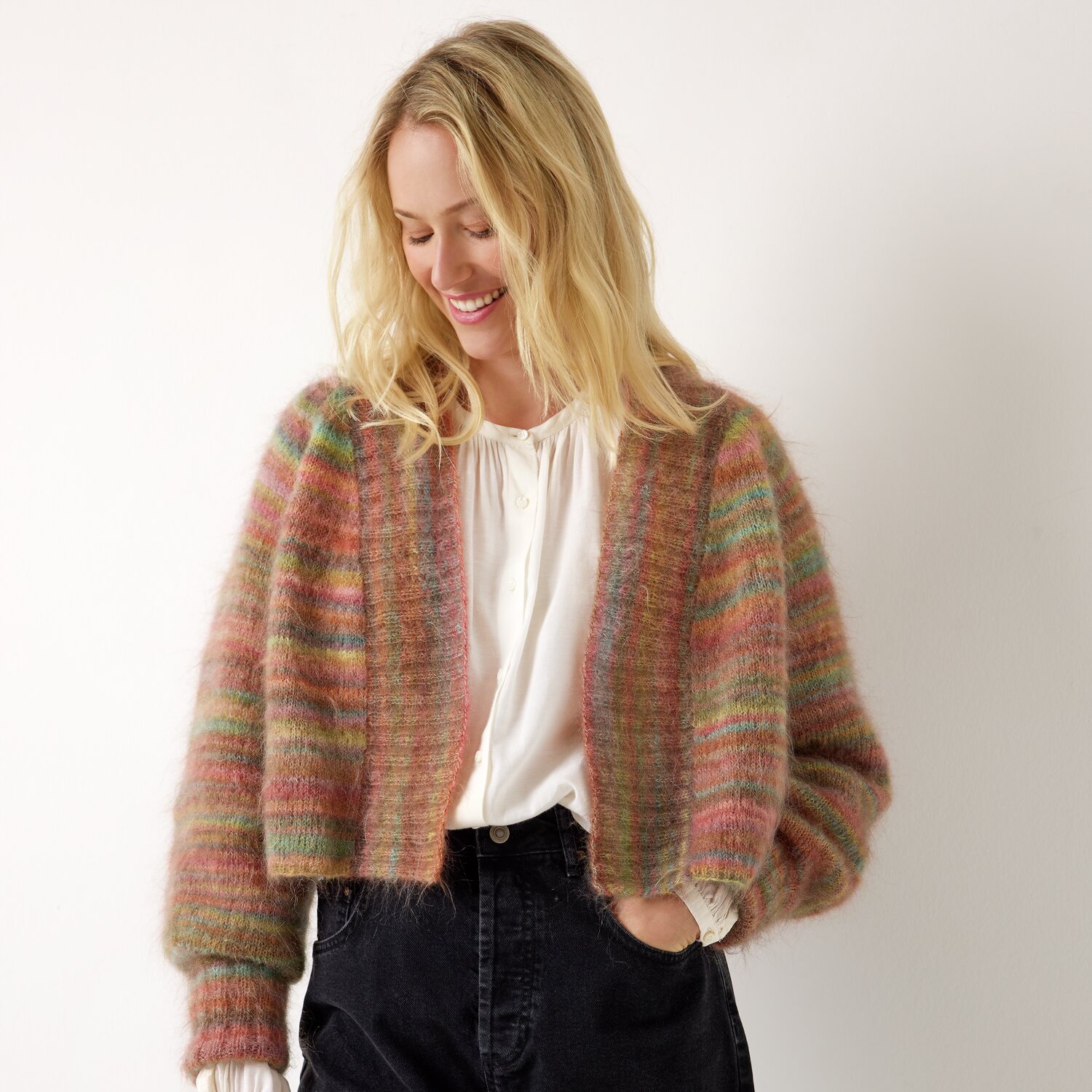 Strickset Jacke Modell 16 aus Die Neue Masche No. 13 von idee. Creativmarkt Strickset Jacke Modell 16 aus Die Neue Masche No. 13: Bunte, gestrickte Jacke in warmen Tönen, getragen von einer lächelnden Frau vor hellem Hintergrund.