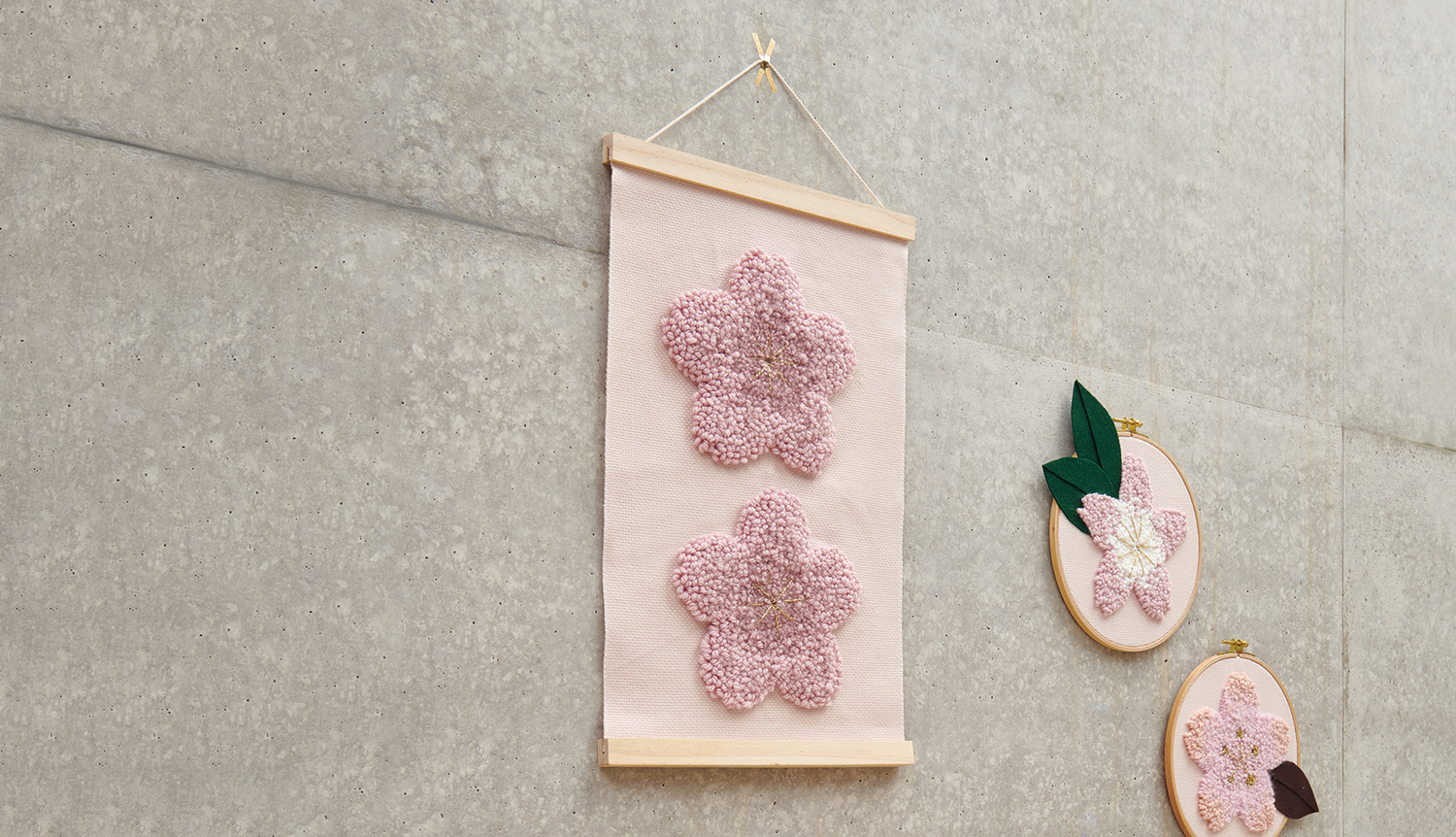 Anleitung Punch Needle Wandbehang mit rosa Blüten zeigt zwei rosa Blumen auf beigem Stoff, aufgehängt an einer grauen Wand.
