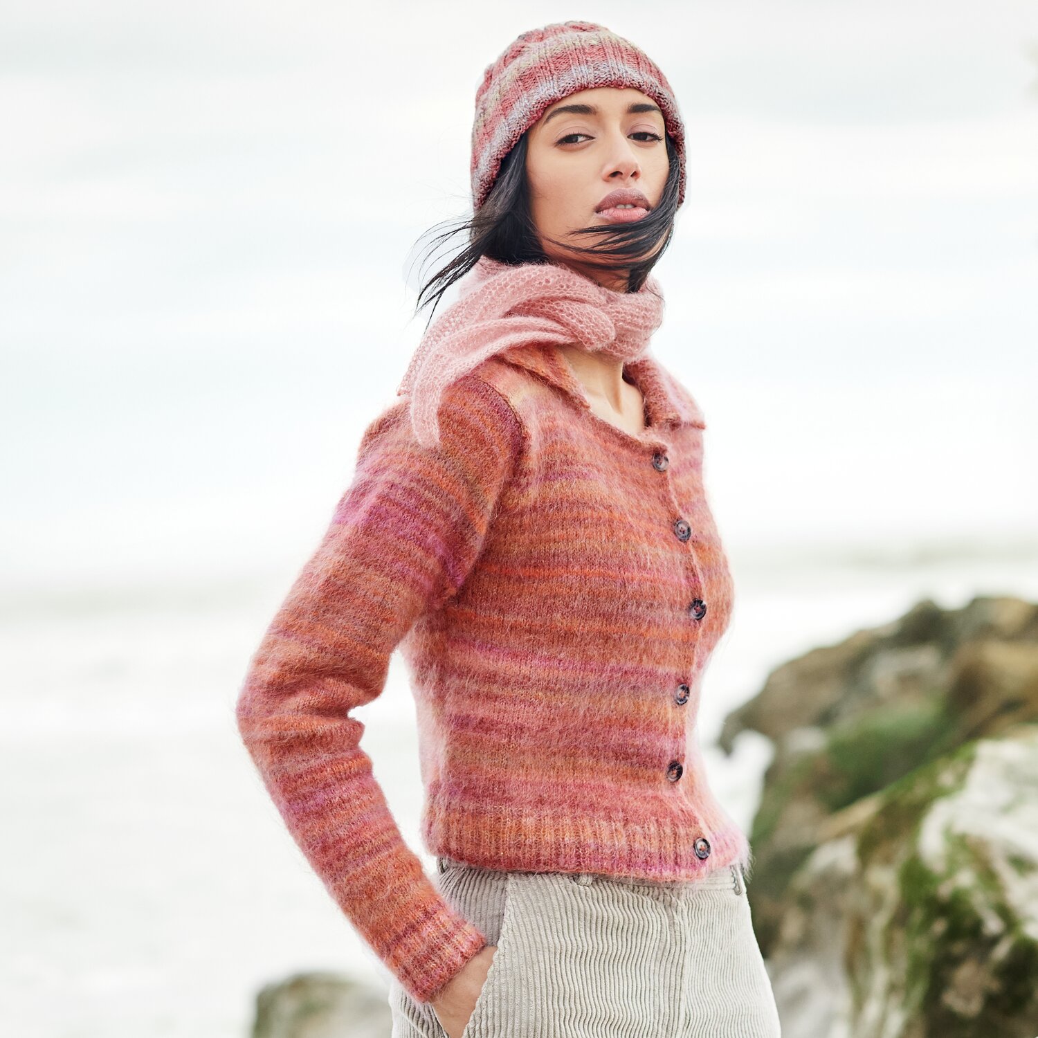 Strickset Minituch Modell 41 aus Lovewool No. 21 von Rico Design Strickset Minituch Modell 41: Frau in rosa Strickjacke und Mütze am Strand, Lovewool No. 21.