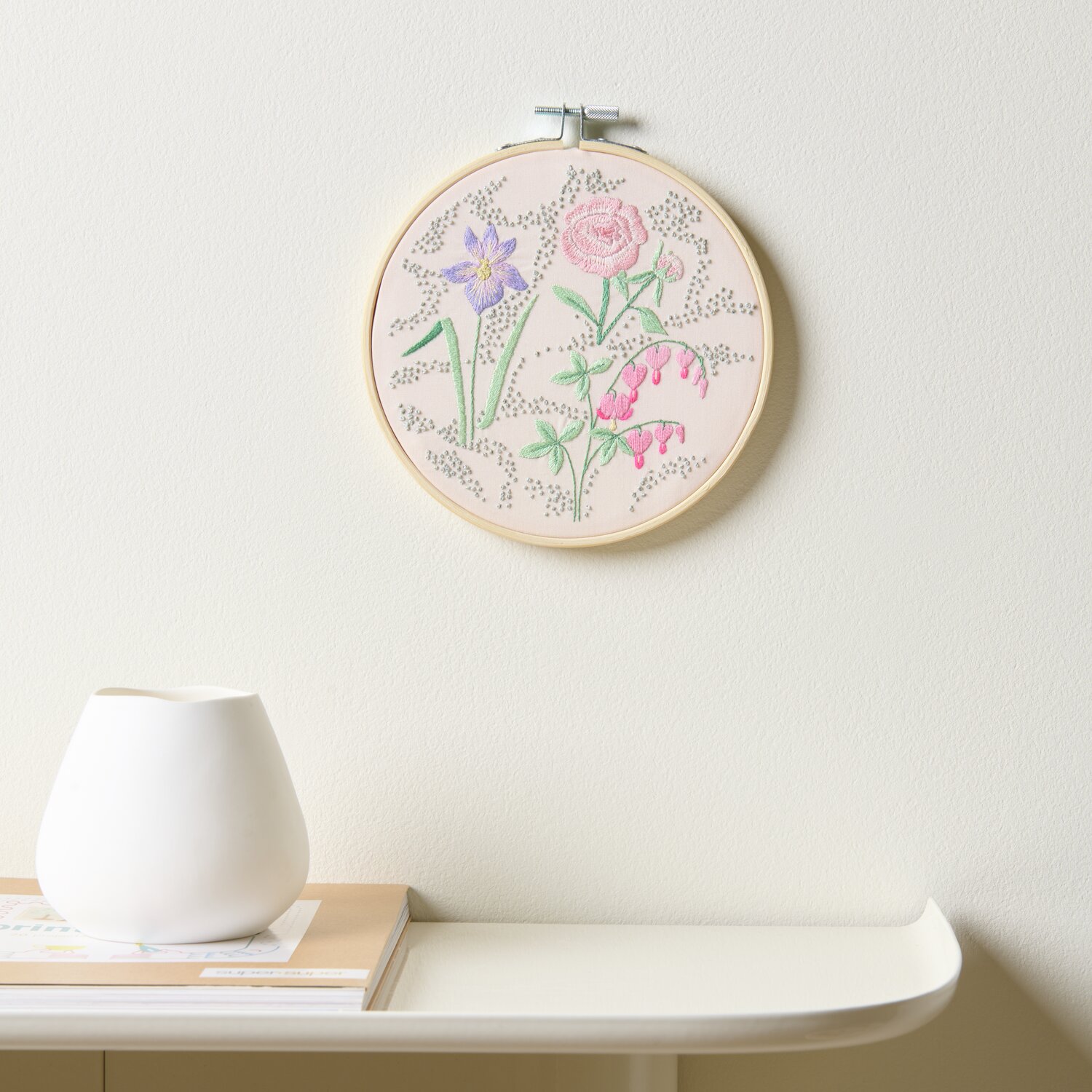 Stickpackung Vorgezeichnet Frühlingsblumen: Runde Stickerei mit bunten Blumen auf beigem Stoff, Ø15cm, an einer weißen Wand hängend.