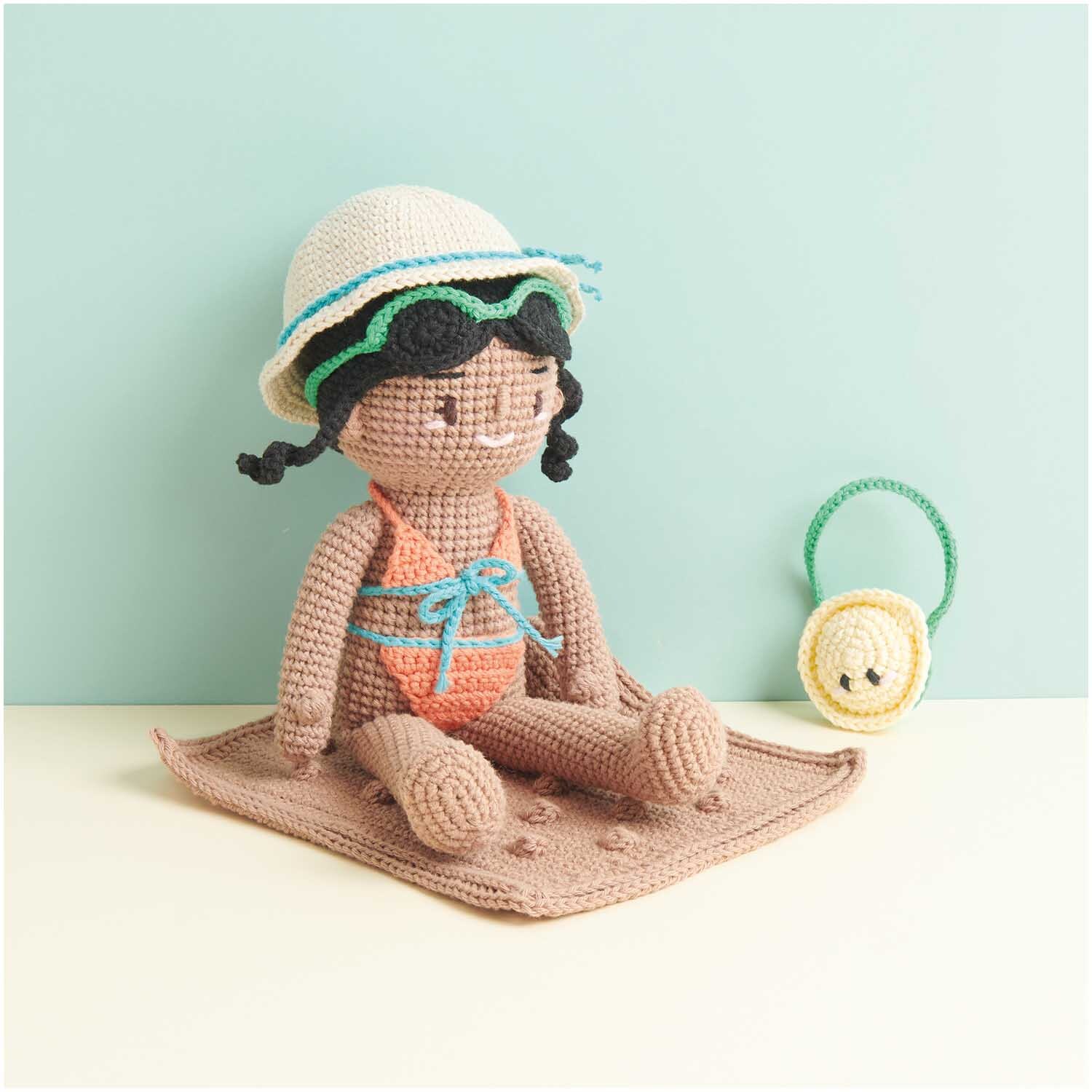 Creative Ricorumi dk: Gehäkelte Puppe in Badeanzug auf Decke, mit Hut und Tasche, in sanften Farben.