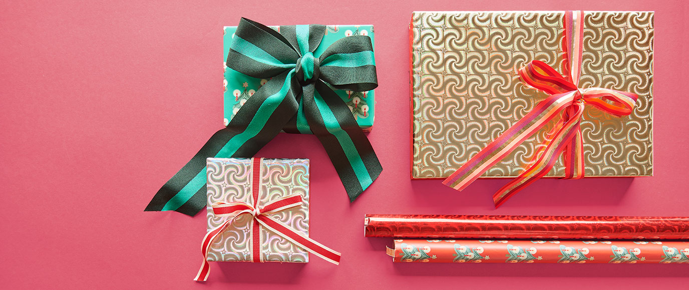 Drei Geschenke in unterschiedlichem Papier und Schleifen verpackt, daneben zwei Rollen Geschenkpapier. Flatlay von Produkten.