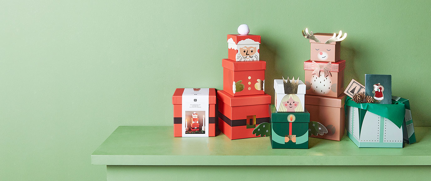Geschenkschachteln jeweils so übereinandergestapelt, dass sie einen Weihnachtsmann, ein Rentier und einen Engel darstellen. Fotosetting grüne Wand.