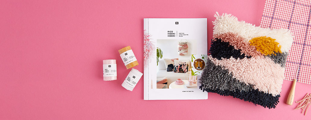 Ein geknüpftes Kissen, ein Knüpf-Heft, verschiedenes Knüpfgarn, Knüpfstramin und ein Knüpfhaken. Flatlay von Produkten.