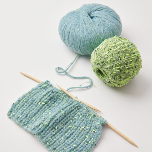 Ein Strickstück an zwei Stricknadeln und ein blaues Garn, sowie ein grünes Beilaufgarn.