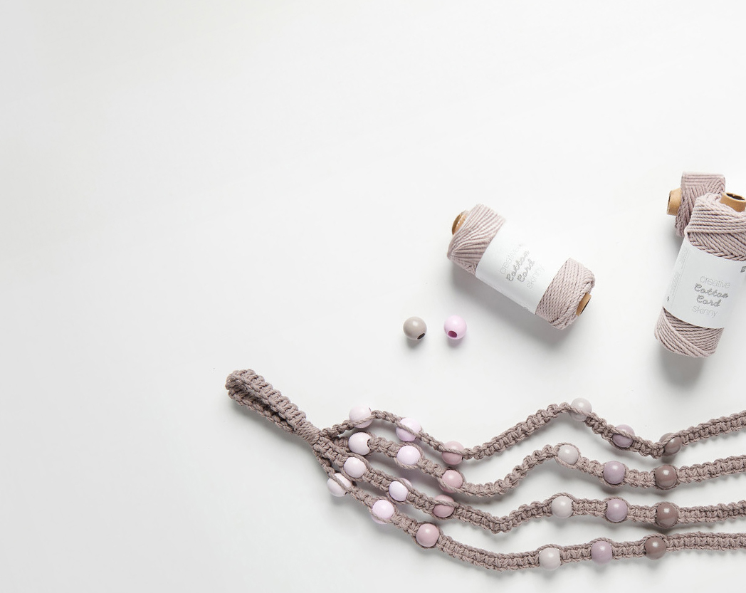 Bräunliches Kordelgarn mit Holzkugeln verknotet, sowie drei verpackte Kordelgarne. Flatlay von Produkten.