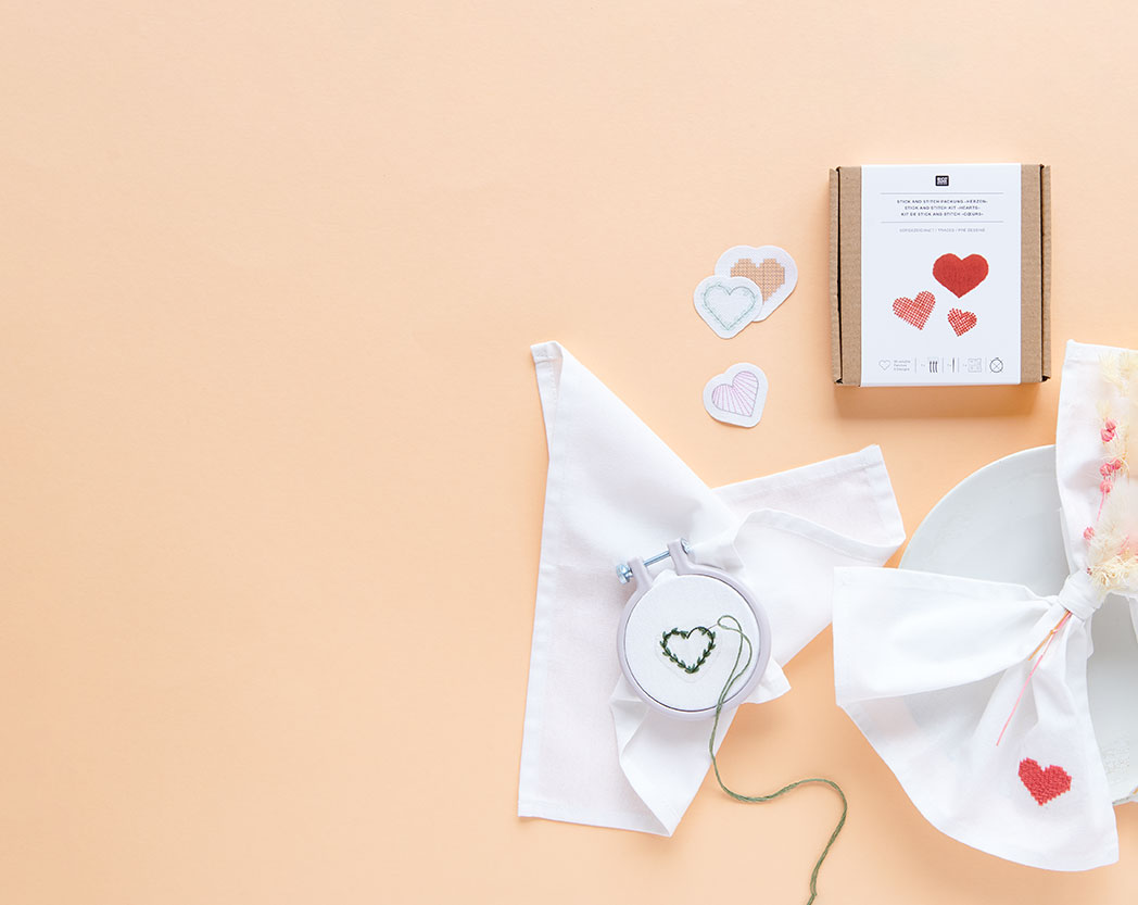 Zwei Stick and Stich Packungen mit Stickvorlagen, daneben eine bestickte Serviette und eine Serviette in einem Stickring mit Nadel. Flatlay von Produkten.