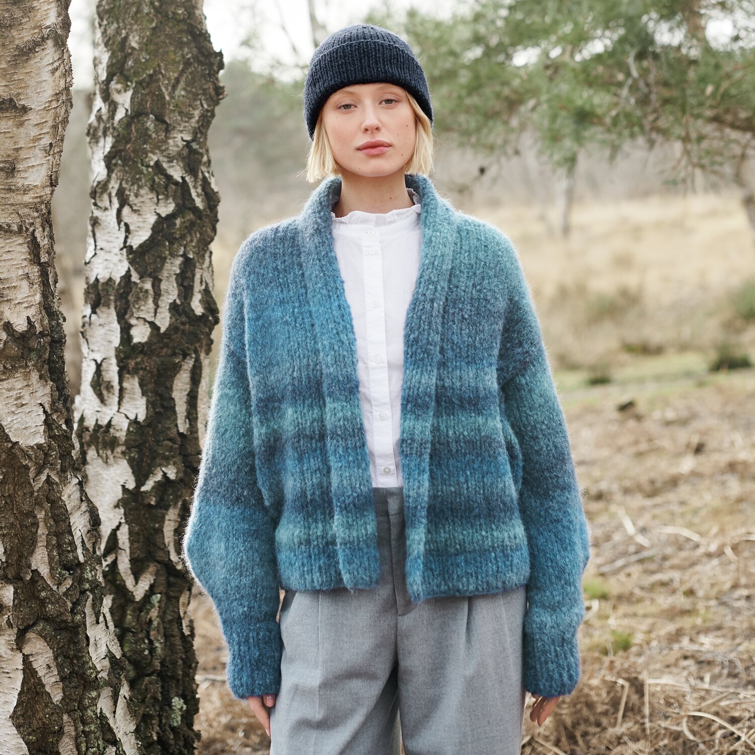 Strickset Cardigan Modell 15b aus Lovewool No. 21 von Rico Design Strickset Cardigan Modell 15b aus Lovewool No. 21: Frau trägt blauen Cardigan im Freien, kombiniert mit grauer Hose und Mütze.