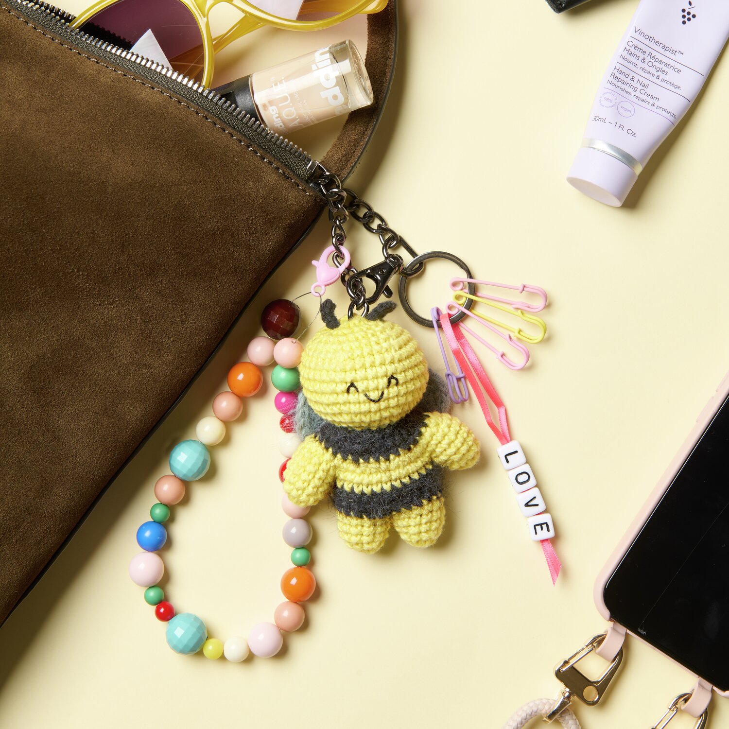 Ricorumi Rico Chingus Schlüsselanhänger in Pastellgelb mit bunten Perlen und LOVE-Schriftzug auf einem Tisch neben einer Tasche.