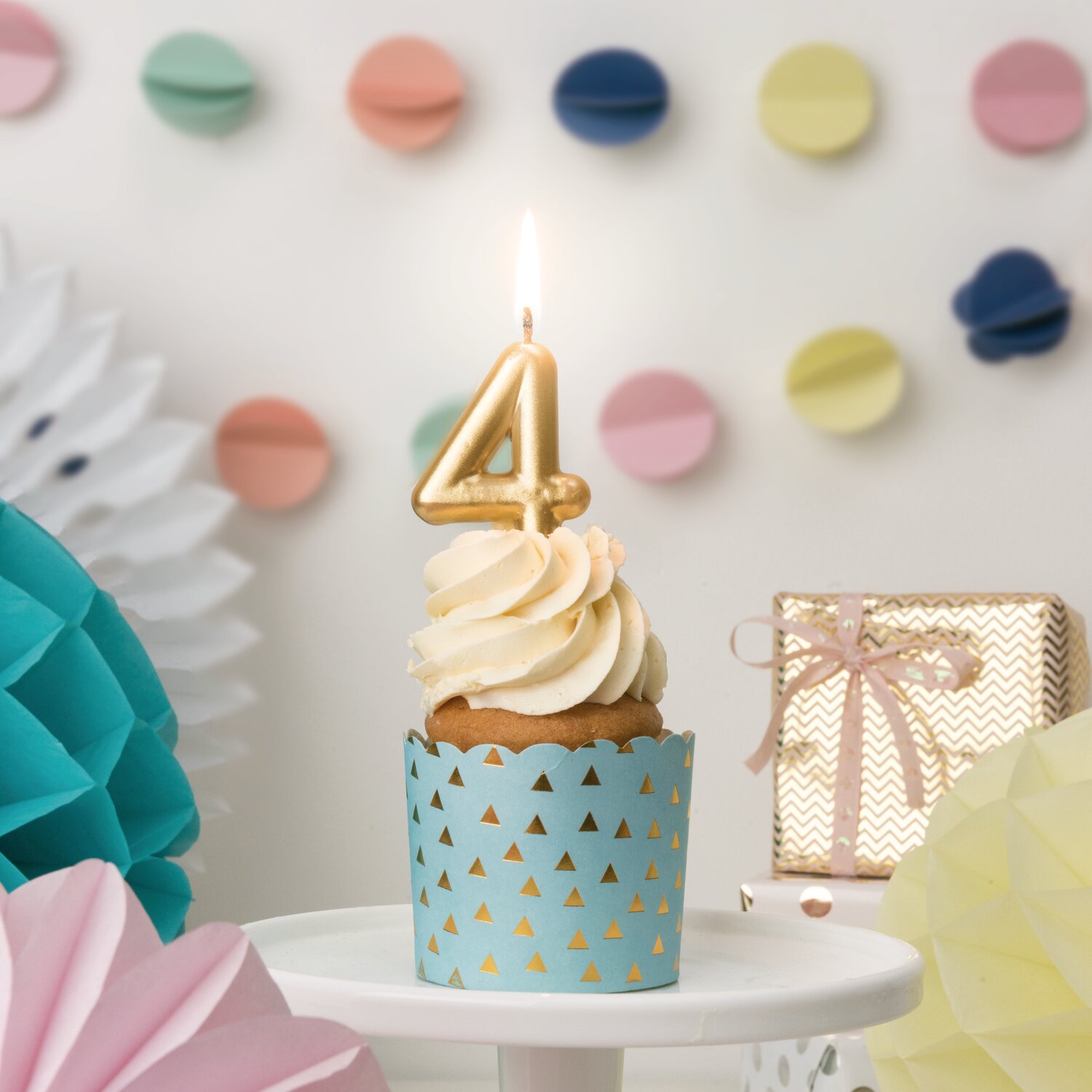 Zahlenkerze Gold von Rico Design auf brennender Cupcake-Kerze, inszeniert vor pastellfarbener Partydeko mit Geschenkbox und Papierornamenten.