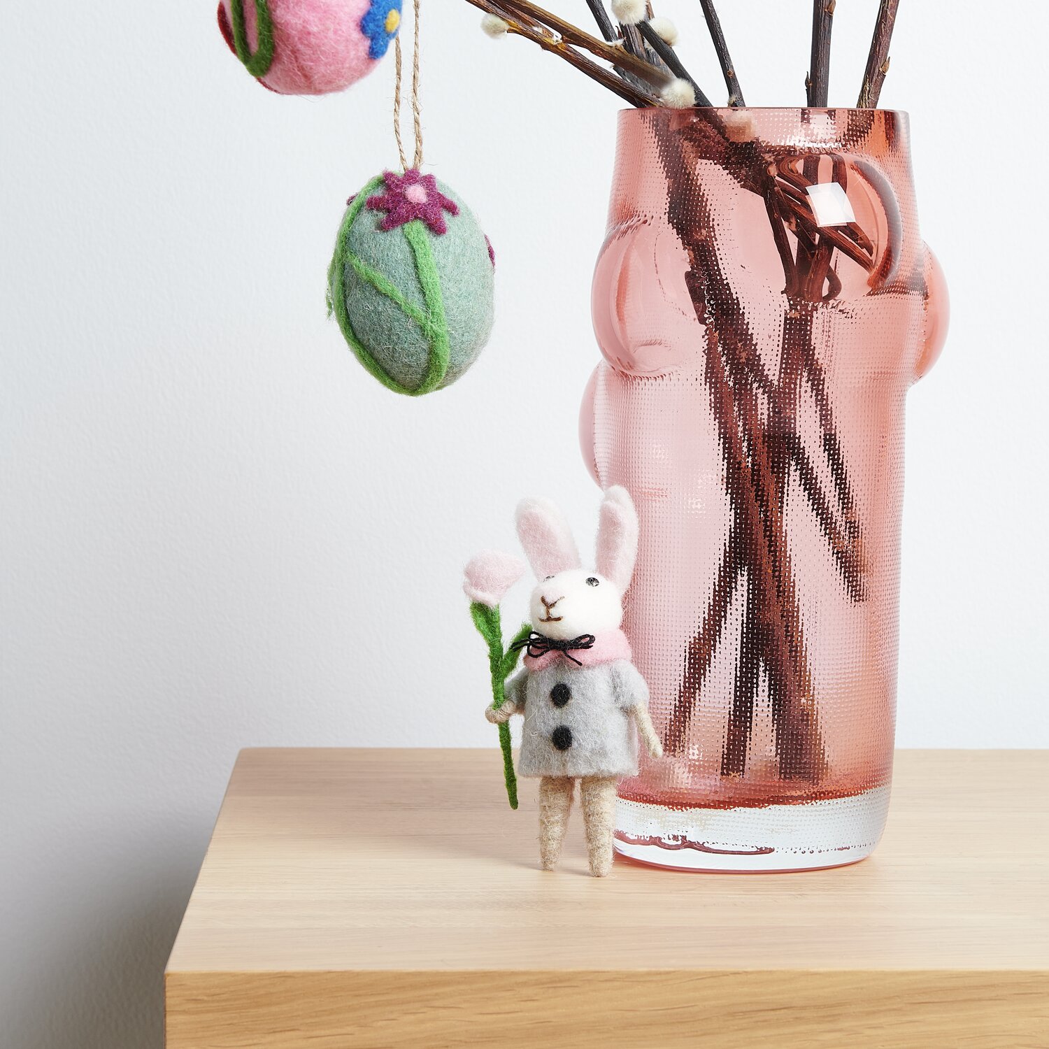 Aufhänger Filz-Ei Purpur-Blume und Filz-Hase mit Jacke vor rosa Vase auf Holzoberfläche.