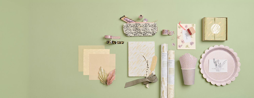 Flach ausgelegte Papeterie- und Geschenkartikel in Pastellfarben auf hellgrünem Hintergrund, darunter Notizbuch, Geschenkverpackungen, Karten, Geschenkpapier und dekorative Accessoires.