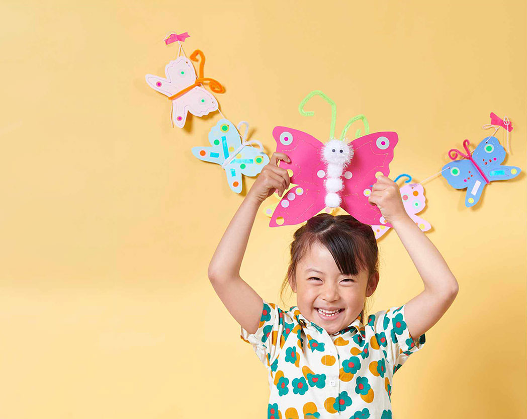 Ein Mädchen hält einen pinken gebastelten Schmetterling über ihrem Kopf, im Hintergrund hängt eine Girlande aus bunten gebastelten Schmetterlingen.