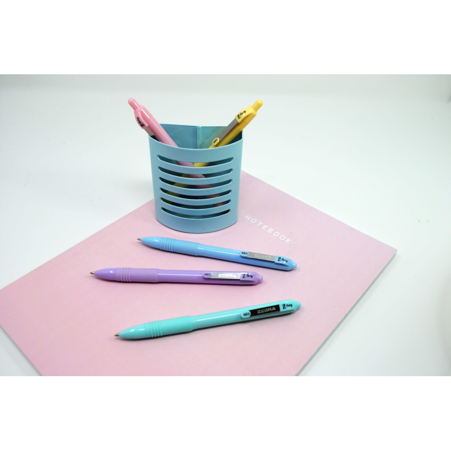 Kugelschreiber Z-Grip Pastell auf rosa Notizbuch, daneben ein blauer Stiftehalter mit weiteren Stiften.