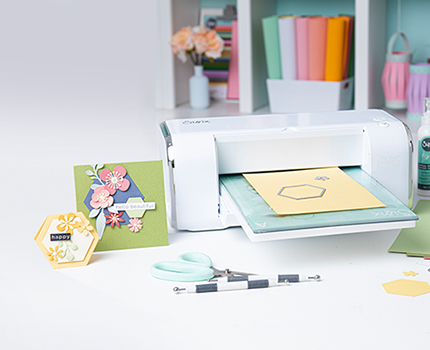 Stanzmaschine mit gelbem Papier und Framelit, sowie verschiedene Thinlits und Modellierzubehör, daneben gebastelte Karten mit Blumen-Design aufgestellt.
