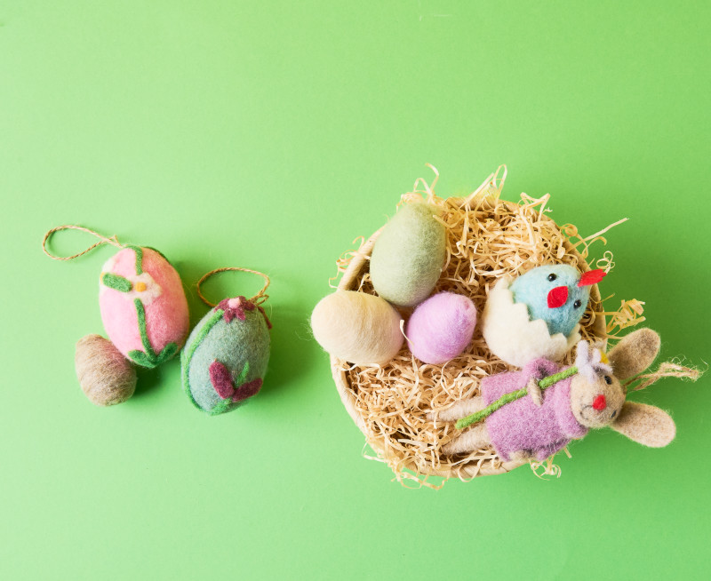 Ein Osterkorb gefüllt mit Ostergras, gefilzten Eiern und gefilzten Osterfiguren, daneben drei weitere gefilzte Eier. Flatlay von Produkten.