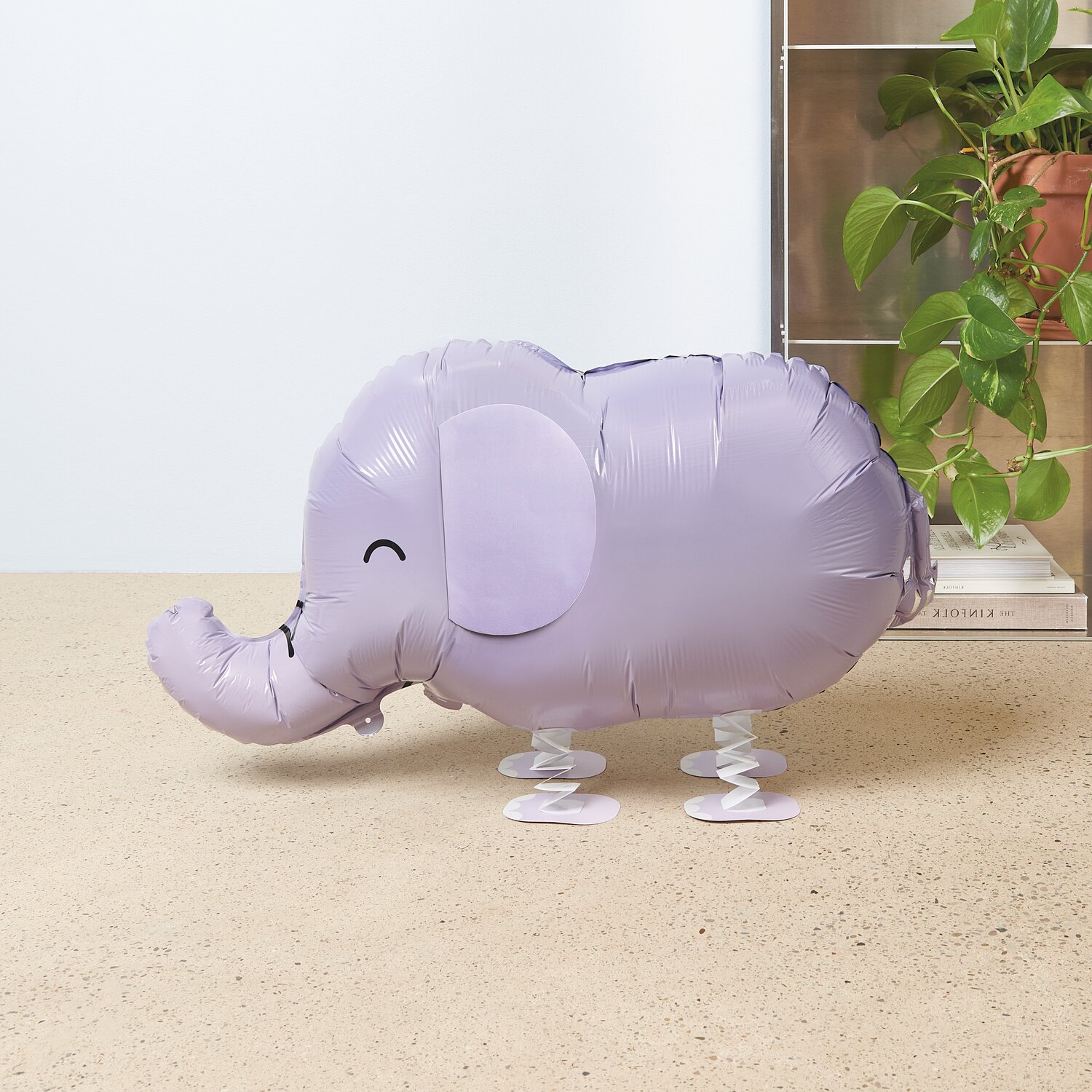 Folienballon Airwalker Elefant in Lila auf hellem Tisch, dekorativ mit grüner Pflanze im Hintergrund.