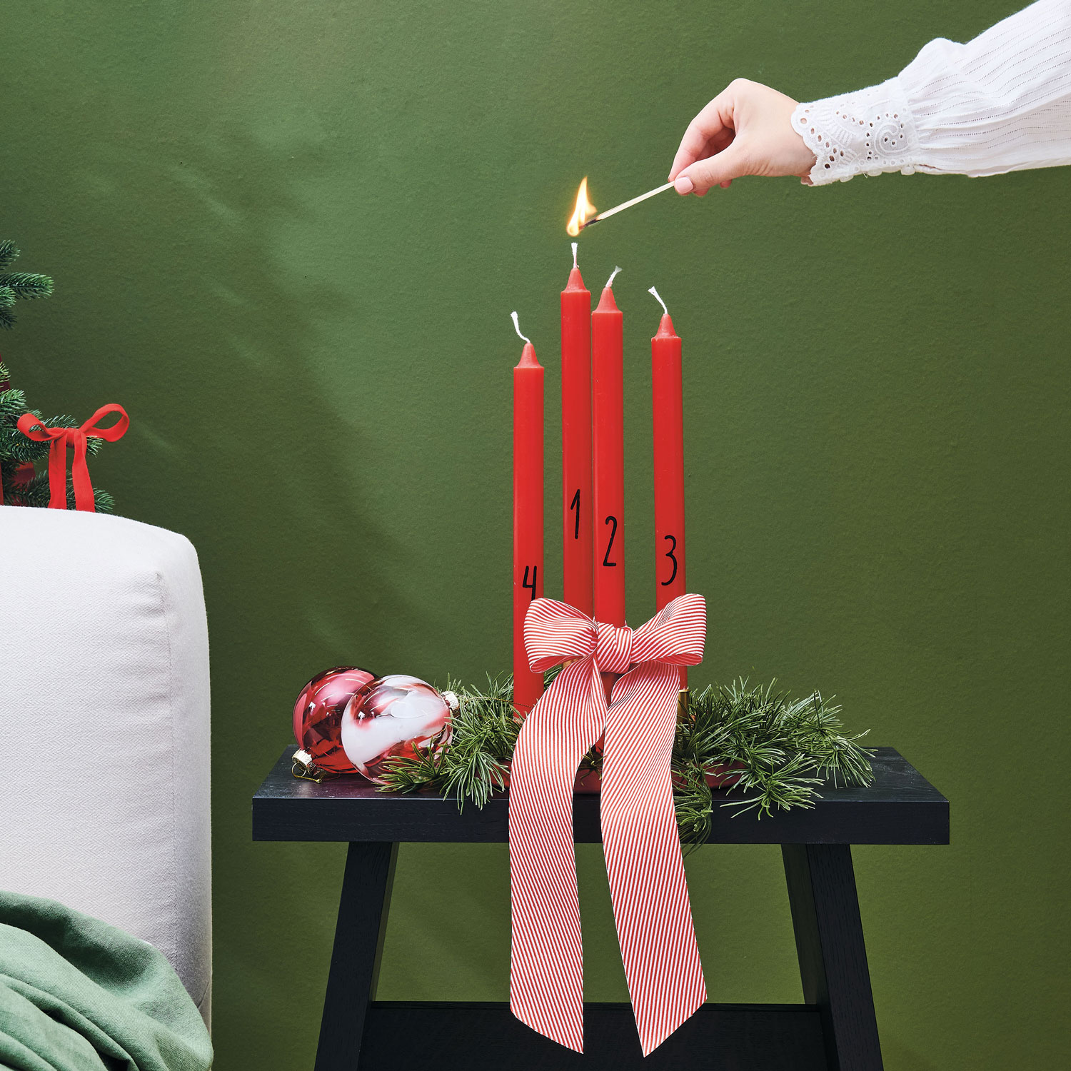 Ein Adventskranz mit roten Stabkerzen, welche mit einem Streichholz angezündet werden, einer roten Schleife und Weihnachtsbaumkugeln. Fotosetting grüne Wand.