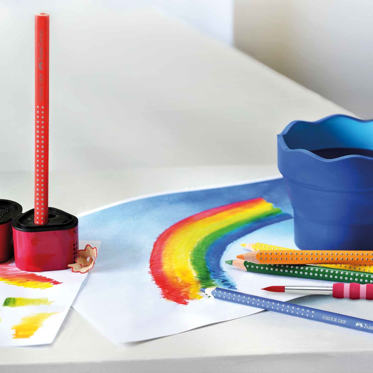 Colour Grip 2001 Buntstifte von Faber Castell auf einem Tisch mit Regenbogenzeichnung und Farbtöpfen.