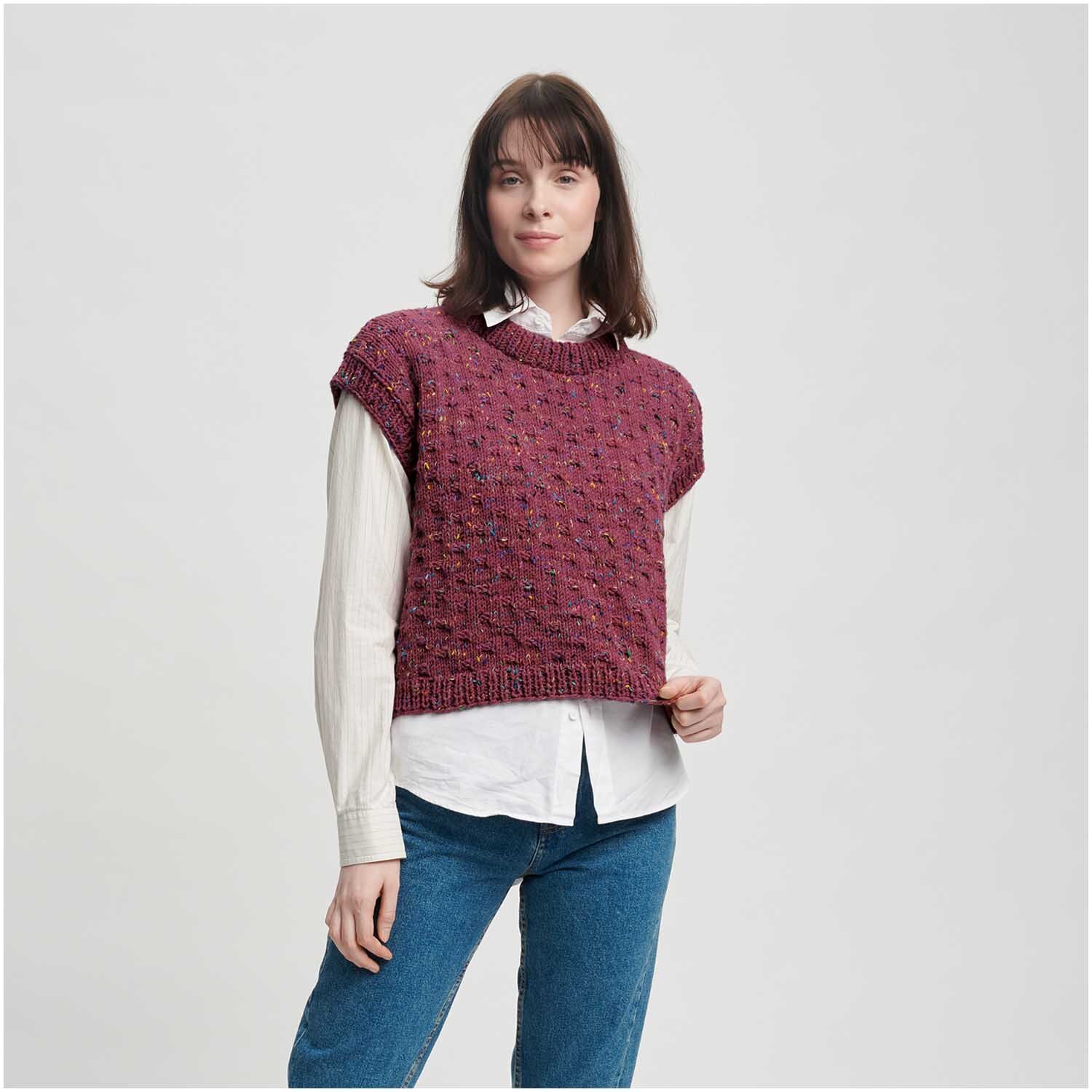 Classik: Frau trägt gestricktes, ärmelloses Oberteil in Mauve von Wolle Rödel, kombiniert mit weißer Bluse und blauen Jeans.